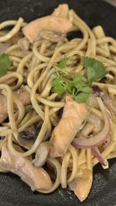 מנת אלפרדו אסייאתי סלמון של הג'פניקה עם פסטה מתחי סלמון טרי וכוסברה על גבי צלחת שחורה