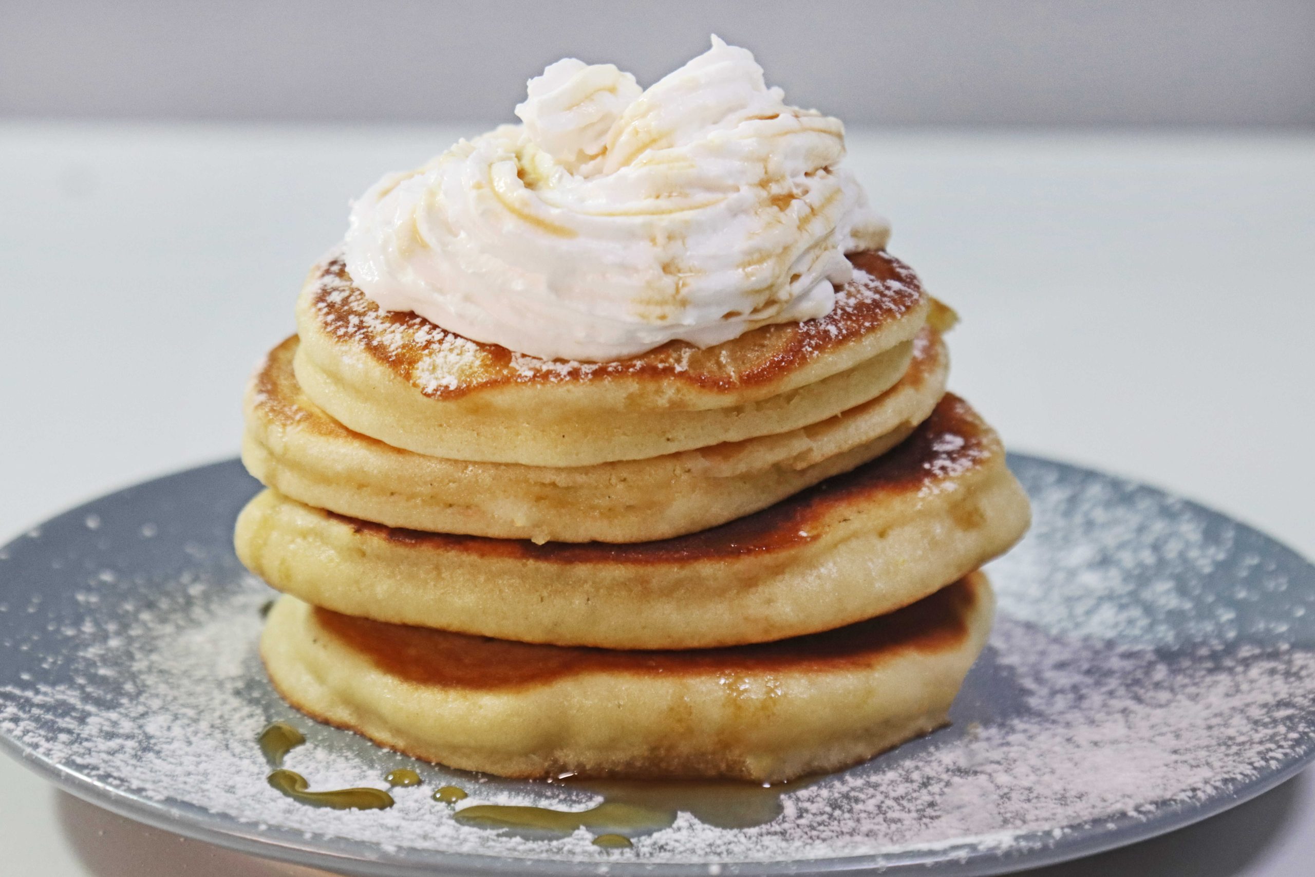 ערמת פנקייקים אווריריים עם גרידת תפוז על גבי צלחת אפורה עם קצפת מעל וזילוף סירופ מייפל