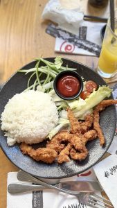 ארוחת אצבעות עוף - ג'פניקידס - אורז לבן עם רצועות עוף, סלט וקטשופ על גבי צלחת שחורה על שולחן עץ בהיר